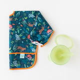 Bumkins Sleeved Bib: Jungle lays folded next to a green transparent bowl & spoon set on a white background.