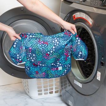 A person holds a Bumkins Sleeved Bib 2 Pack: Jungle and Animal Print in front of an open front-loading washing machine.