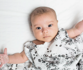 A baby wrapped in a Lulujo Inc. Bamboo Swaddle, Black Floral lies on a white blanket, gazing directly at the camera wide-eyed.