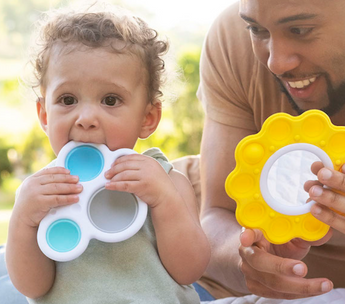 A toddler chews a Dimpl Billow Bright by Fat Brain Toy Co, while an adult holds a yellow silicone bubble pop toy, enjoying the sunny day.