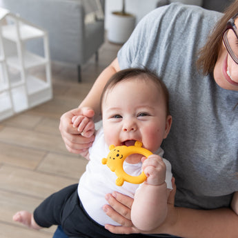 A baby smiles on a woman's lap, happily chewing a Bumkins Silicone Flat Teether: Winnie the Pooh.