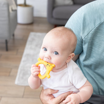 A blue-eyed baby enjoys the Bumkins Silicone Flat Teether: Winnie the Pooh, soothing gums while snuggled on a lap indoors.