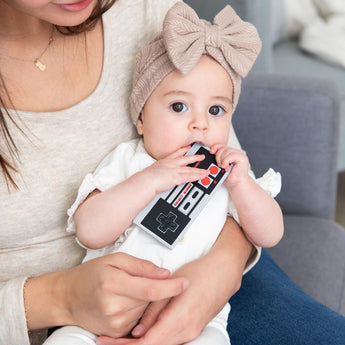 A baby in a large beige bow chews a Bumkins Silicone Teether: NES™ Controller while held by a woman on a gray couch.
