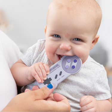 A smiling child in a gray shirt chews a video game controller-shaped teether held by a hand with blue nail polish.