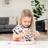 A blonde girl in a black & white bib eats a meal from a Bumkins Silicone Grip Tray: Pink with pink utensils at a white table.