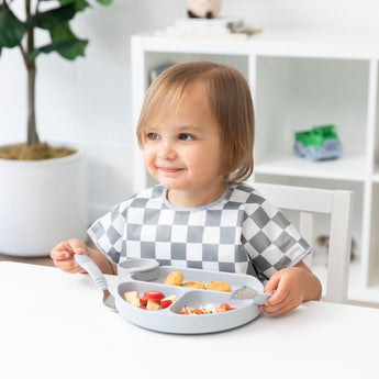 A toddler in a Bumkins Junior Bib: Charcoal Check smiles, eating food from a gray divided plate at a white table.
