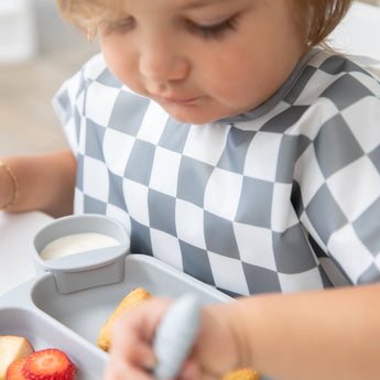 A toddler wearing a Bumkins Junior Bib: Charcoal Check focuses on eating from a divided tray while sitting at a white table.
