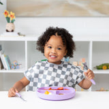 A curly-haired toddler smiles while eating, wearing a Bumkins Junior Bib: Charcoal Check, at a white table with a purple plate of fruit.