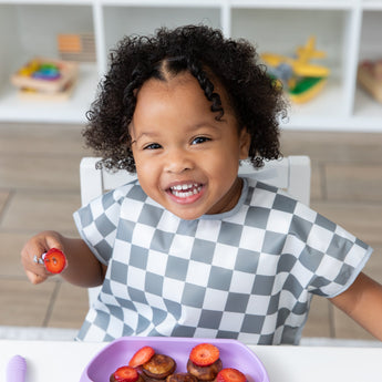 A child smiling, wearing Bumkins Junior Bib: Charcoal Check at a white table while eating pancakes and strawberries from a purple plate.