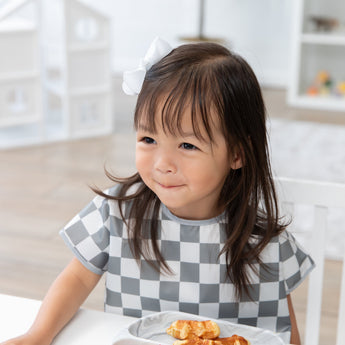 A young girl in a Bumkins Junior Bib: Charcoal Check sits at a table with waffles, smiling softly.