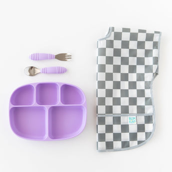 A purple baby plate with matching fork and spoon and and Bumkins Junior Bib: Charcoal Check on a crisp white background.