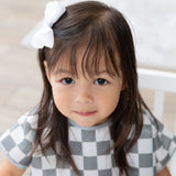 A young girl with brown hair and a bow smiles in a Bumkins Junior Bib: Charcoal Check, made from waterproof fabric.