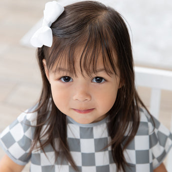 A young girl with brown hair and a bow smiles in a Bumkins Junior Bib: Charcoal Check, made from waterproof fabric.