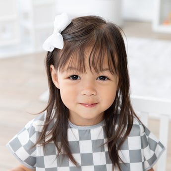 A young girl sits and smiles in a bright room, wearing a Bumkins Junior Bib: Charcoal Check with waterproof, easy-wipe fabric.