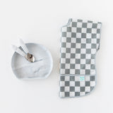 Bumkins Junior Bib: Charcoal Check beside a gray bowl with two spoons on a white background.