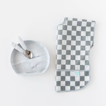 Bumkins Junior Bib: Charcoal Check beside a gray bowl with two spoons on a white background.
