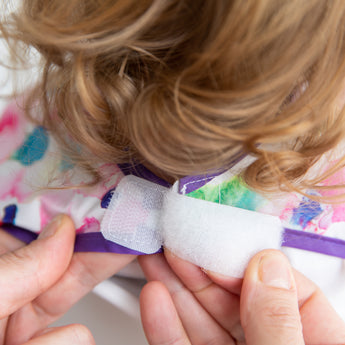 Close-up of curly-haired child wearing a Bumkins Junior Bib: Watercolor, secured with a hook-and-loop fastener by adult hands.