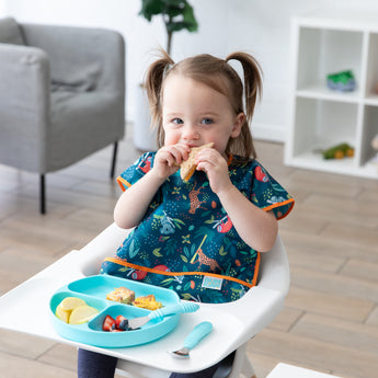 A child in a Bumkins Junior Bib: Jungle enjoying a sandwich from a blue dish seated in a white highchair.