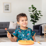 A boy grins, wearing an animal patterned bib as he eats from a Bumkins Silicone Grip Dish: Tangerine with matching utensils.