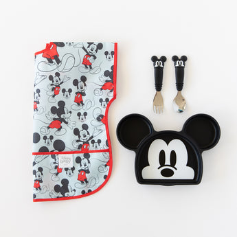 Bumkins Junior Bib: Mickey Mouse Classic lays folded next to a Mickey Mouse-shaped plate and matching utensils on a white background.