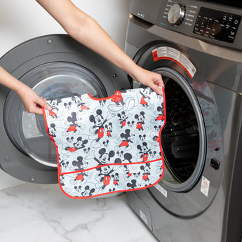 A person holds Bumkins Junior Bib: Mickey Mouse Classic in front of an open front-loading washing machine.