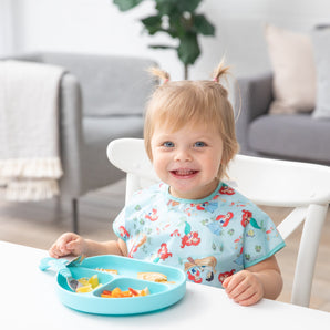 A smiling toddler in pigtails wears a Bumkins Junior Bib: Ariel and eats happily from a divided plate with utensils at a table.