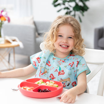 A curly-haired girl smiles, wearing a Bumkins Junior Bib: Ariel, eating fruits and veggies from a sectioned plate at a table.