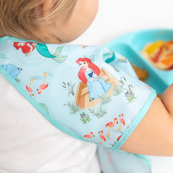 A child dons the Bumkins Junior Bib: Ariel, featuring the red-haired mermaid, next to a blue tray of food.