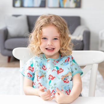 A smiling curly-haired girl at a table, wearing a Bumkins Junior Bib: Ariel with a cozy living room backdrop.
