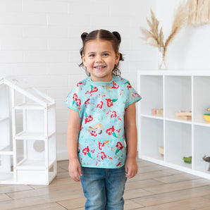 A girl with pigtails wearing a Bumkins Junior Bib: Ariel smiles, standing in front of a dollhouse and toy-filled shelves.