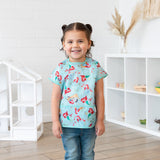 A girl with pigtails wearing a Bumkins Junior Bib: Ariel smiles, standing in front of a dollhouse and toy-filled shelves.