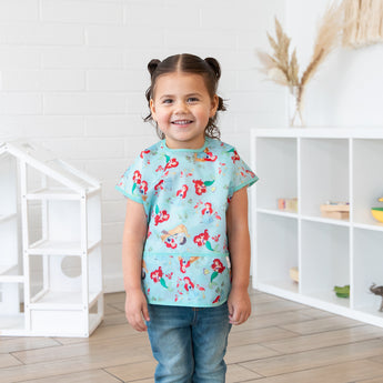 A girl with pigtails wearing a Bumkins Junior Bib: Ariel smiles, standing in front of a dollhouse and toy-filled shelves.