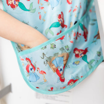 A child places their hand in the pocket of a Bumkins Junior Bib: Ariel featuring colorful cartoon mermaids and flamingos.