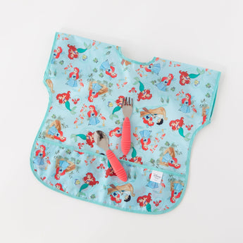 Bumkins Junior Bib: Ariel, featuring mermaid illustrations, lays flat on a white background with a red-handled fork and spoon on top.
