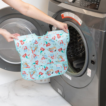 A person holds a Bumkins Junior Bib: Ariel, featuring cartoon designs and waterproof fabric, in front of a front-loading washing machine.