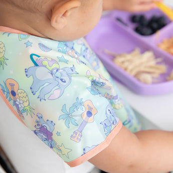 Close-up of a child's shoulder wearing a Bumkins Junior Bib: Disney Aloha Stitch with a purple plate of food in front.