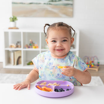 A smiling girl in a Bumkins Junior Bib: Disney Aloha Stitch eats from a purple divided plate in a bright playroom.