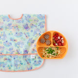 A bib from Bumkins Junior Bib 2 Pack: Stitch Star-ring Up Trouble lays flat next to an orange plate with pasta, fruit, & veggies on white.