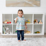 A young girl in a Bumkins Junior Bib: Disney Aloha Stitch stands in front of toy-filled shelves in a bright playroom.