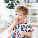 A boy wearing a Bumkins Junior Bib: Super Mario™ & Luigi eats a snack in a bright room with toy filled shelves and a plant.