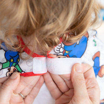 Hands fastening a Velcro strip on a Bumkins Junior Bib featuring Super Mario™ & Luigi with a cartoon-themed collar.