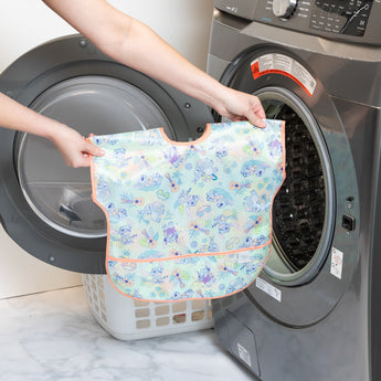 A person holds a Bumkins Junior Bib 2 Pack: Stitch Star-ring Up Trouble in front of an open front-loading washing machine.