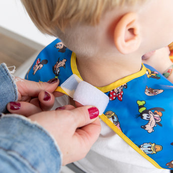 Adult fastening a Bumkins Junior Bib 2 Pack: Mickey & Friends Excellent Eight with Velcro around a young childs neck.