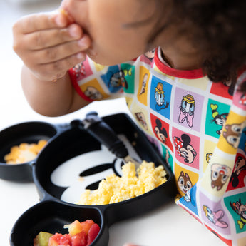 Close-up of a child wearing a bib from Bumkins Junior Bib 2 Pack: Mickey & Friends Excellent Eight eating eggs from a Mickey-shaped plate.
