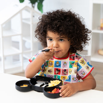 A child in a Bumkins Junior Bib (Mickey & Friends) eats from a black divided plate at a white table.