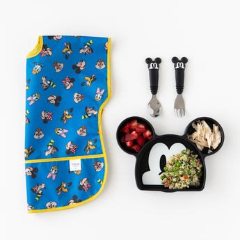 A bib from Bumkins Junior Bib 2 Pack: Mickey & Friends Excellent Eight lays folded next to a Mickey-shaped plate & utensils on white.