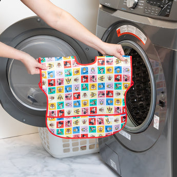 Hands placing a Bumkins Junior Bib 2 Pack: Mickey & Friends Excellent Eight into a front-loading washing machine beside a laundry basket.