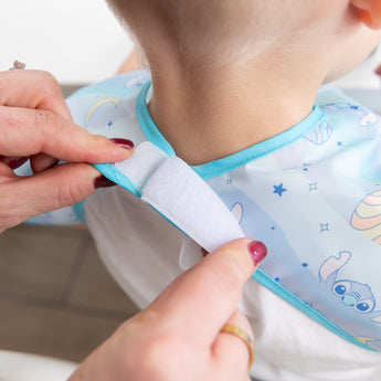 Adult hands fasten the hook-and-loop closure of a Bumkins Junior Bib 2 Pack: Stitch Star-ring Up Trouble around a child's neck.