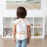 A child in a Bumkins Short-Sleeved Smock: Wander faces a white shelf filled with toys and books, showcasing the neck and waist closures.