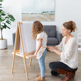 In a cozy room with wall art and plants, a woman adjusts a Bumkins Short-Sleeved Smock: Wander on a girl ready to paint at her easel.